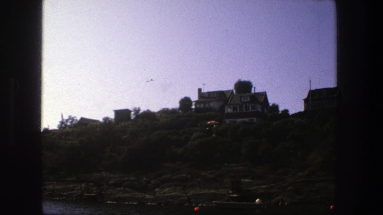 Rocky shoreline below hillside houses, seen through dark film-frame edges.