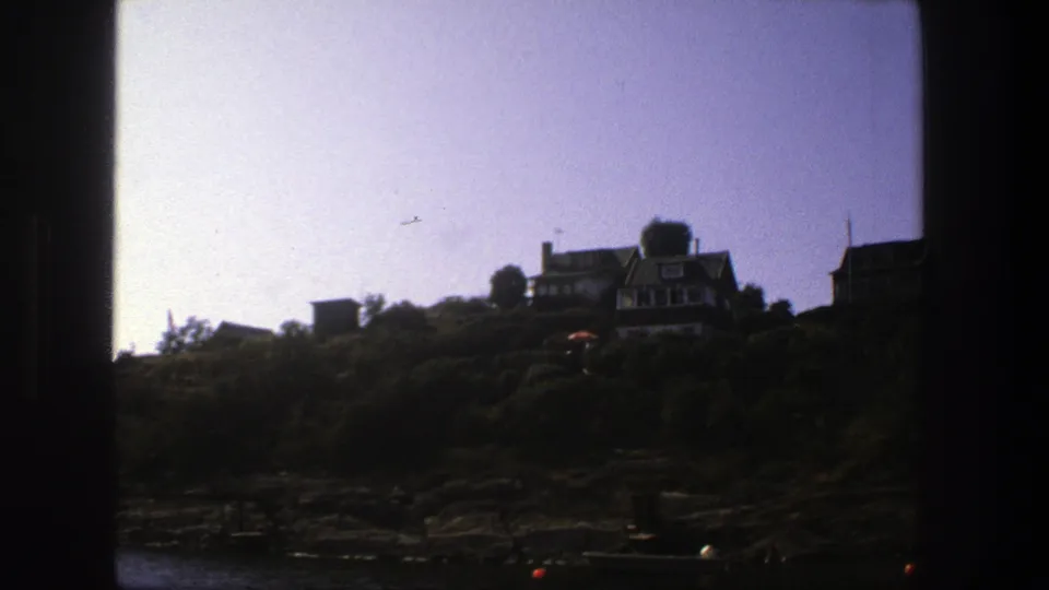 1975 8mm view near Stockholm showing hillside houses above a rocky shoreline with trees, pale sky, and dark frame edges.