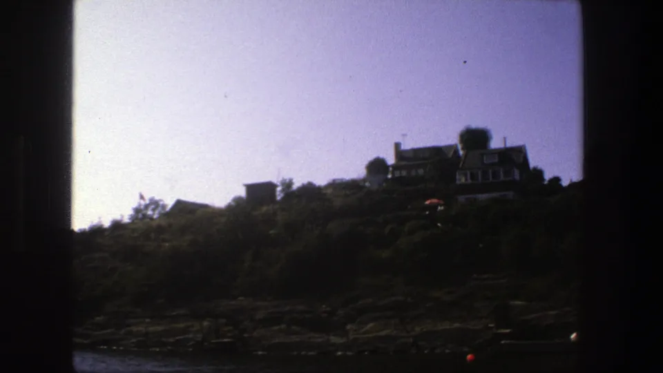 Slow 8mm pass along a Stockholm shoreline, with homes on the hill, dense greenery, rocks, and a faint red buoy near the water.