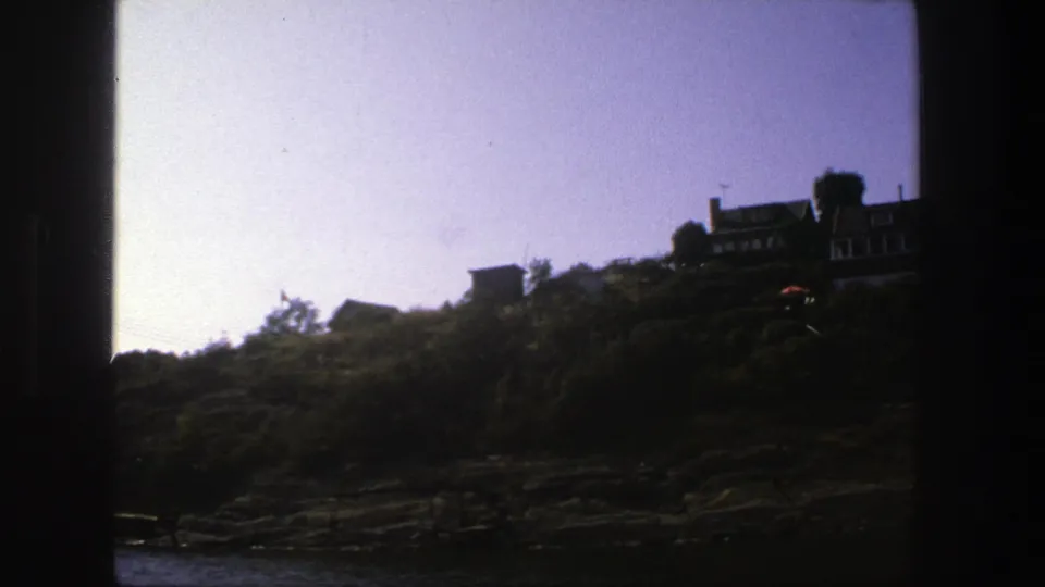 1975 home-movie frame from Stockholm with a dark vertical edge, rocky shoreline, green slope, and houses partly crossing the top of the hill.