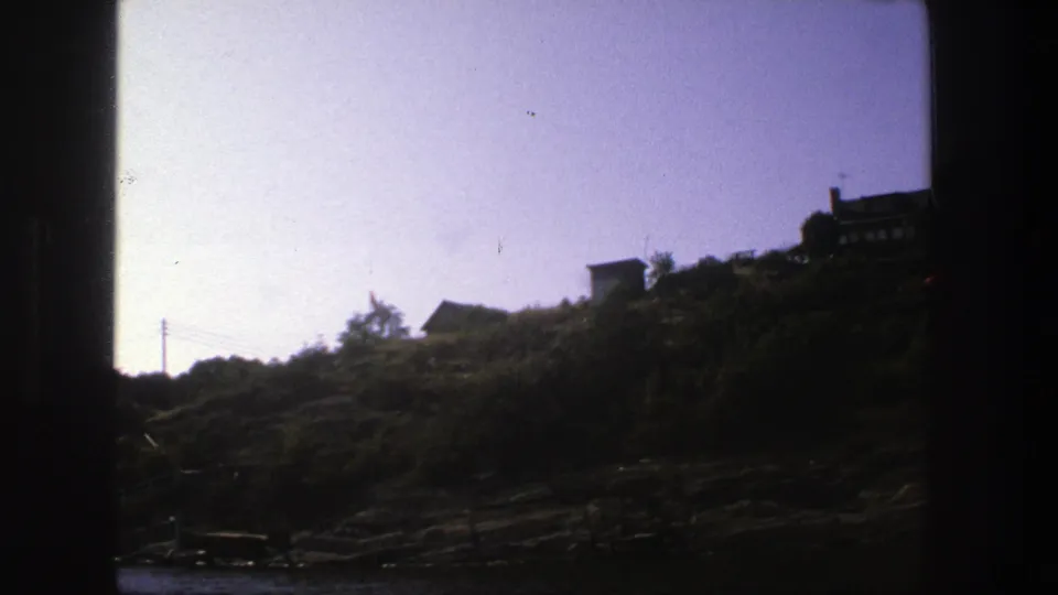 Late frame of the 1975 Stockholm shoreline pass, showing rocks, shrubs, pale sky, a distant house cluster, and dark scan framing.