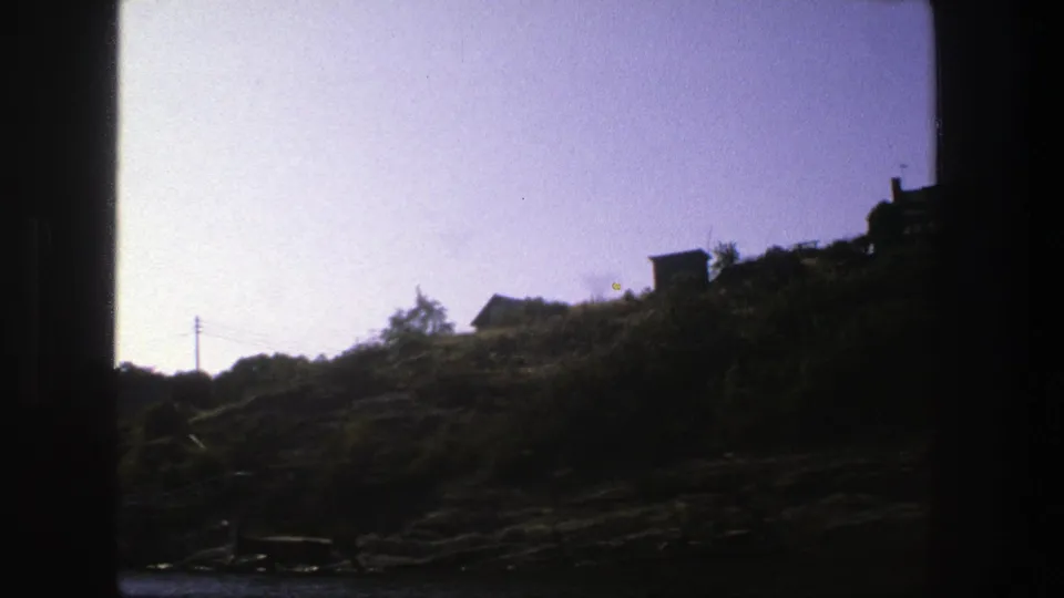 1975 Stockholm 8mm frame showing a shaded rocky hillside, small houses, utility lines, pale sky, and water at the bottom edge.