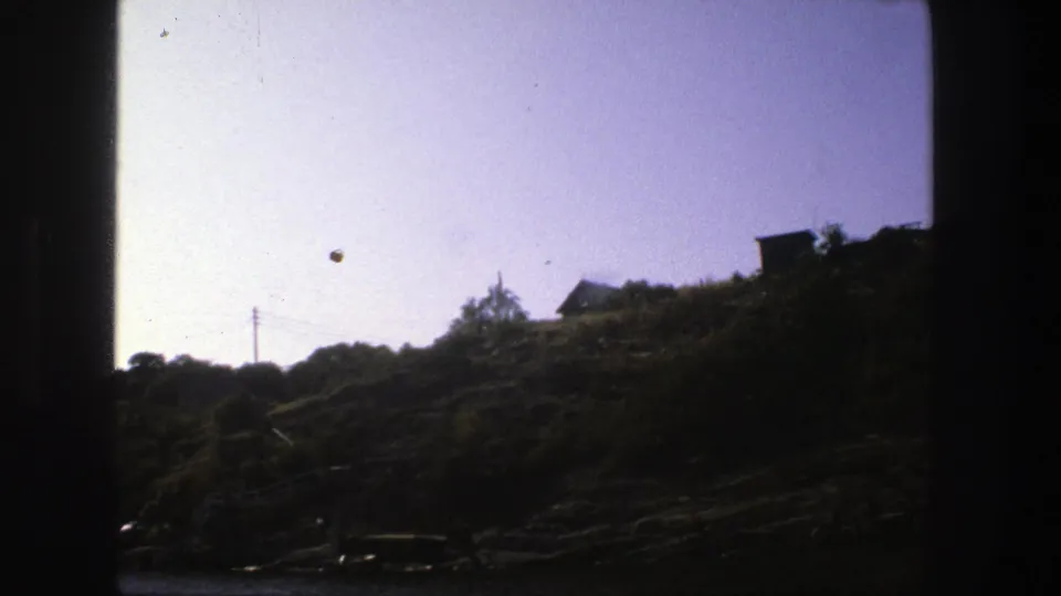 1975 Stockholm 8mm frame of a hillside residential slope, with wires crossing the sky and deep shadow across the rocks near water.