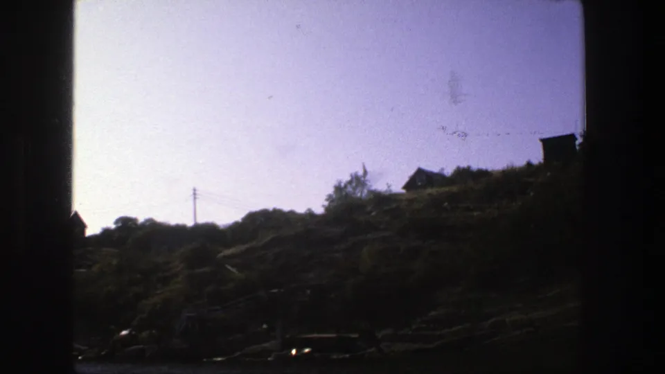 1975 Stockholm home-movie frame showing a backlit rocky shoreline slope with small homes, vegetation, a utility pole, and hazy sky.
