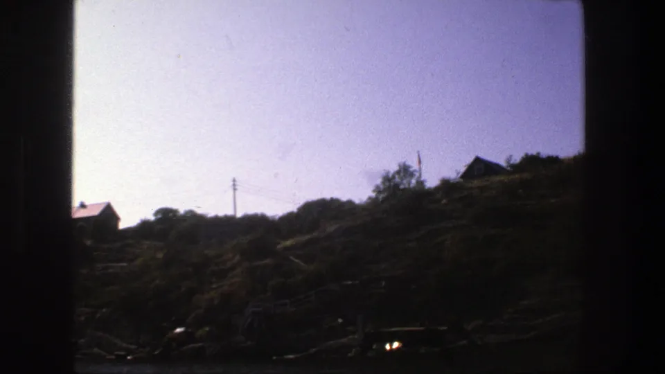 1975 Stockholm 8mm frame with a rocky, vegetated shoreline, silhouetted hillside houses, utility wires, and dark exposure around the water.