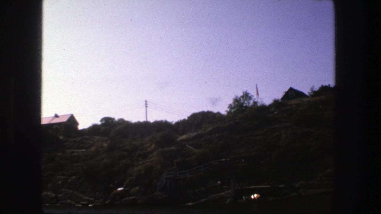 Red-roofed house on a rocky hillside with bushes, trees, utility pole, wires, and pale sky.