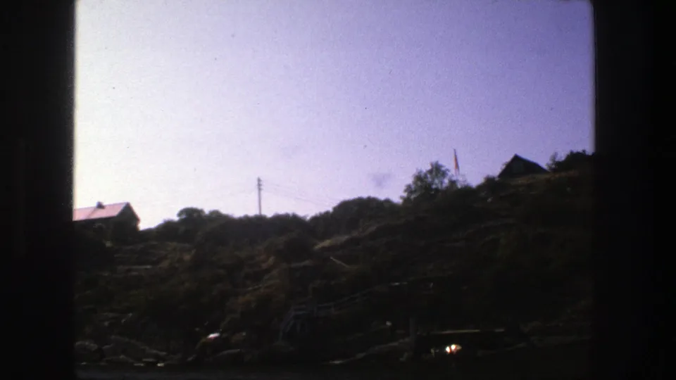 1975 Stockholm 8mm frame showing a red-roofed hillside house among dark shrubs, trees, rocks, utility wires, and pale sky.