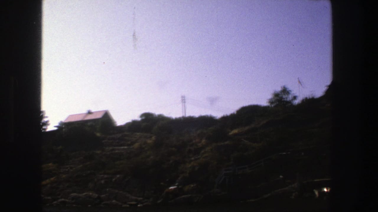 1975 Stockholm home-movie frame with a small red-roofed house on a sloped, rocky hillside beneath utility wires.
