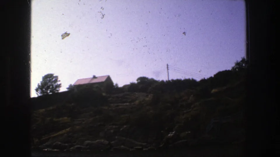 1975 Stockholm 8mm view of a red-roofed house, trees, overhead wires, and a rocky hillside with dark vegetation.