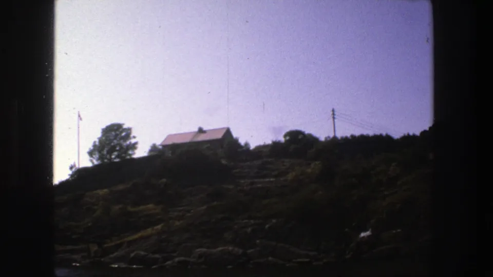 1975 Stockholm frame showing a hillside house near trees, a flagpole, utility wires, rocks, and dense bushes.