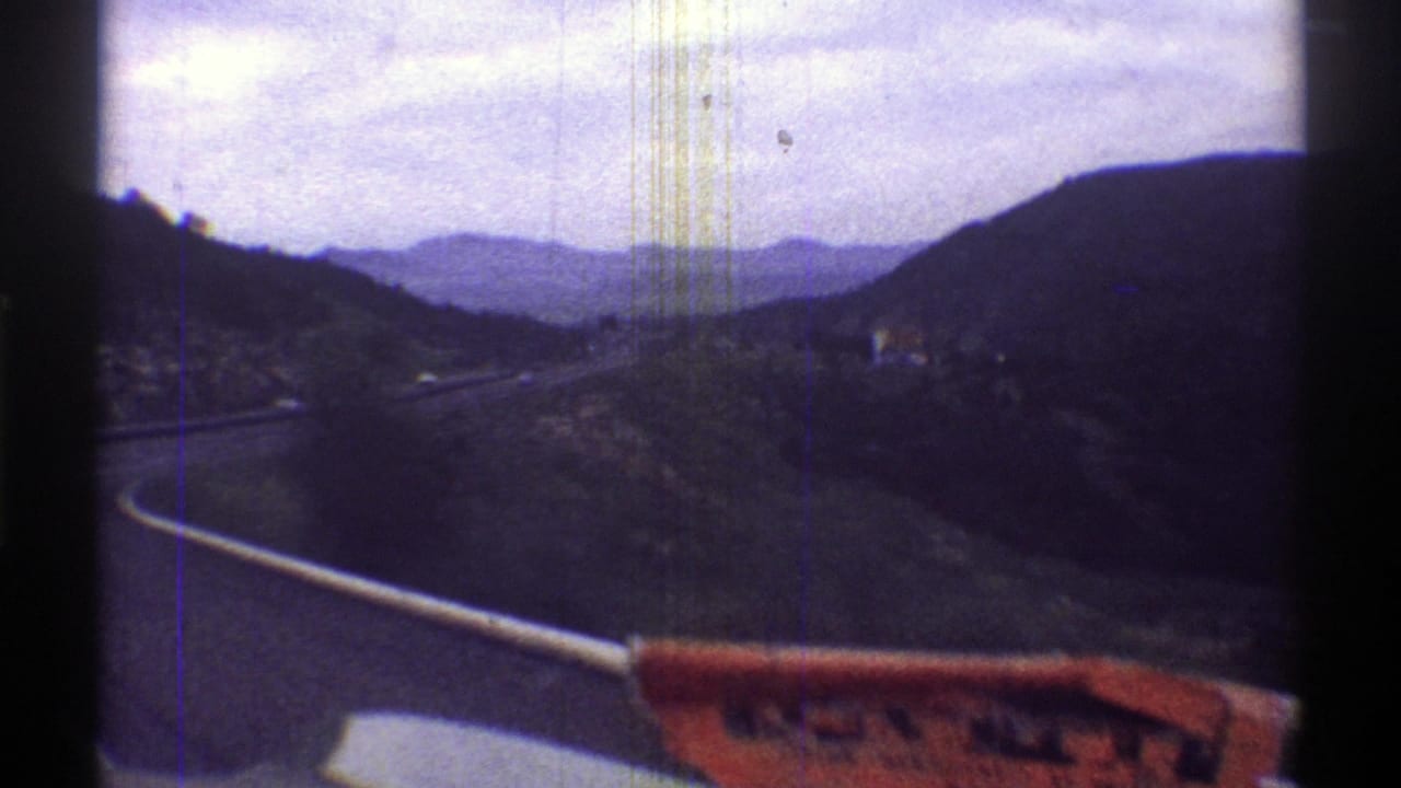 Mountain road curves through a valley with dark hills and a red object near the lower edge.