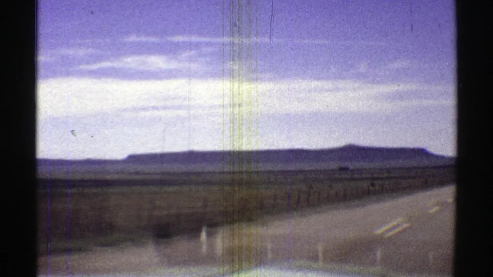 Passenger-view 8mm footage shows a two-lane Arizona road, roadside fence, flat desert, and low mesa formations in the distance.