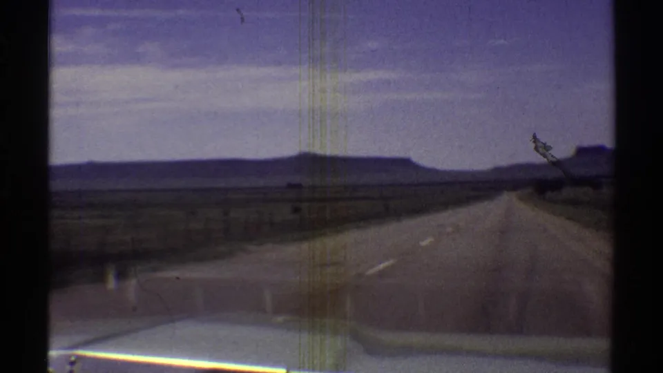 1969 Arizona road-trip view from inside or near a car, showing the highway ahead, open desert, distant mesas, and the car hood.