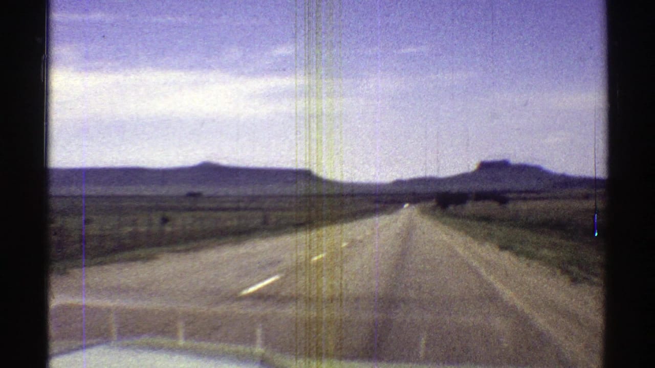 Car hood and windshield view of a straight desert road toward distant mesas under a pale sky.