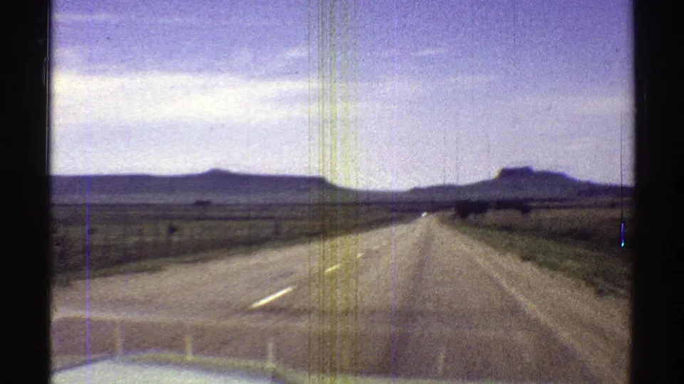 1969 Arizona 8mm windshield view down a straight open road with low grassland, distant flat-topped mesas, and pale clouds.