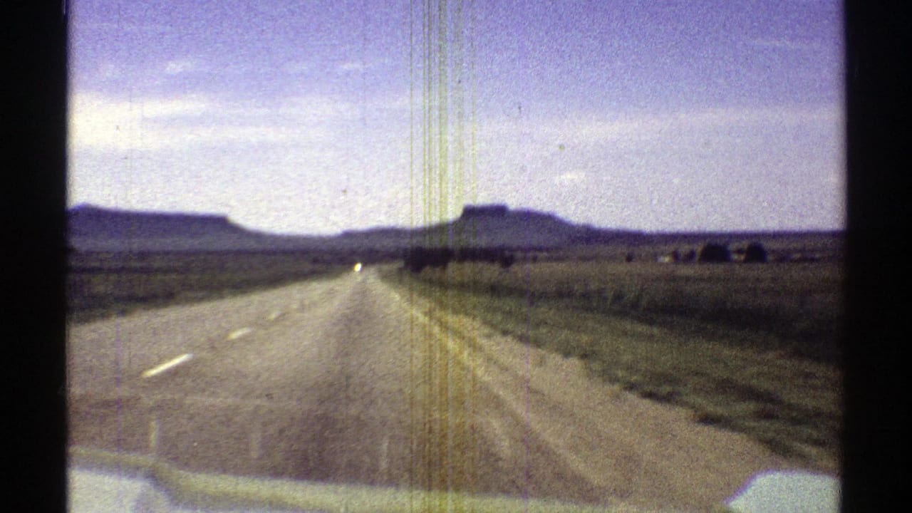 Forward windshield view of an open road crossing grassland with mesas on the horizon.