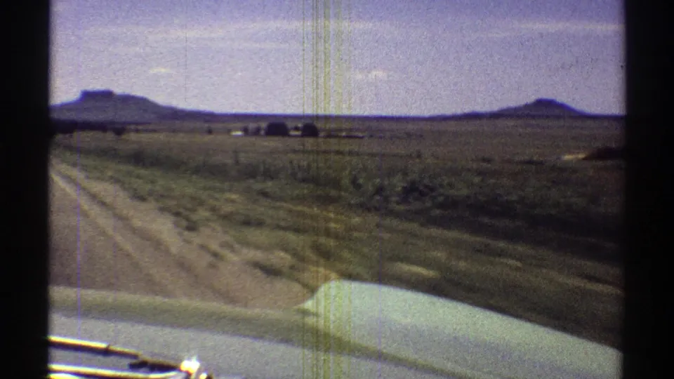 1969 Arizona 8mm frame from a moving car showing roadside grassland, distant mesas, pale sky, and the vehicle hood at the bottom.
