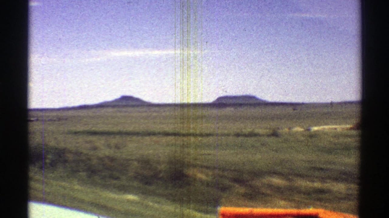1969 Arizona 8mm frame showing open desert terrain, distant mesas, blue sky, and the edge of a car in the foreground.