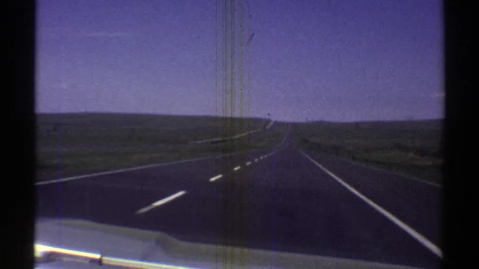 1969 Arizona road-trip frame with a long paved highway, lane markings, low desert terrain, and blue sky seen from inside a moving car.