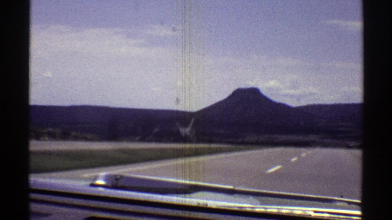 1969 Arizona road-trip frame with the windshield edge and car hood framing a broad highway, roadside grass, and a dark mountain ahead.