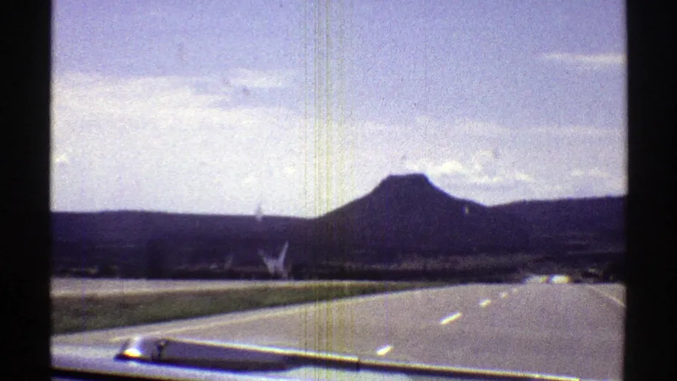 Faded 8mm frame from an Arizona drive, showing a highway, grassy roadside, distant hills, and a flat-topped mountain beyond the windshield.