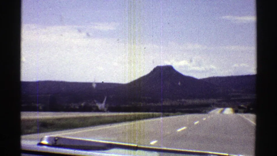 1969 Arizona 8mm road-trip frame showing a quiet highway, car hood, open roadside, distant hills, and a broad mountain under a lightly clouded sky.