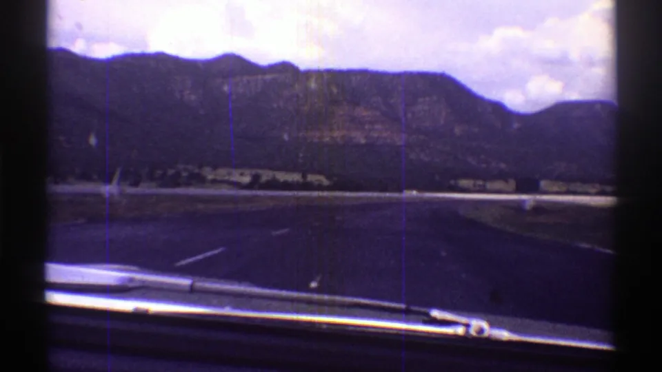 Windshield-level 8mm view from a moving car on an Arizona highway, with mountains and clouds ahead.