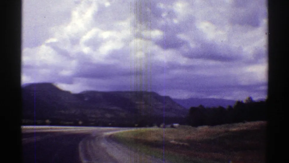 1969 8mm Arizona driving view showing a curved road, roadside trees, mountains, and a cloud-filled sky.