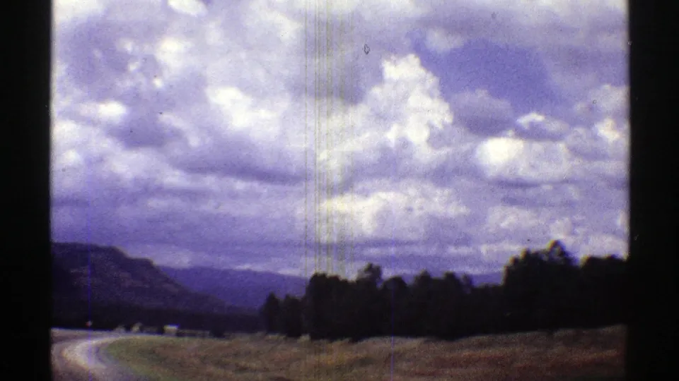 Arizona home-movie frame with a winding road, tree line, layered mountains, and large grey-white clouds.