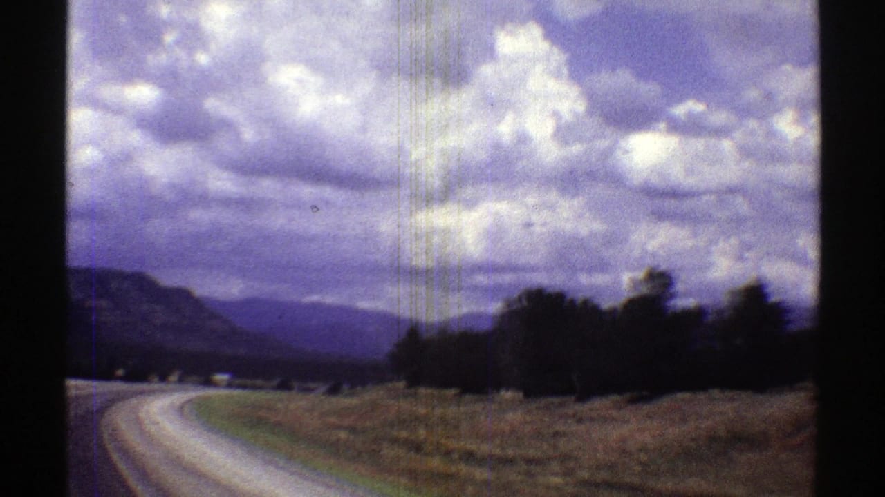1969 Arizona 8mm road-trip frame showing a curving roadside, scrubland, distant mountains, and a dramatic clouded sky.
