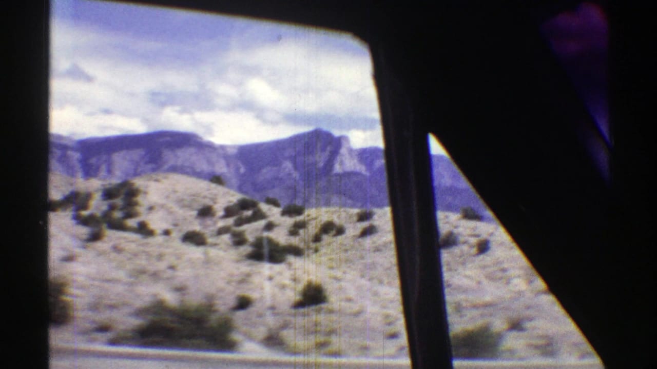 Arizona desert hills and mountain ridges pass by through the dark frame of a moving vehicle window in 8mm footage.