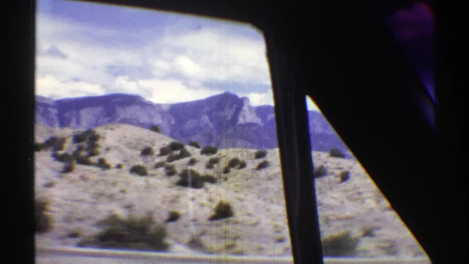 Arizona desert hills and mountain ridges pass by through the dark frame of a moving vehicle window in 8mm footage.