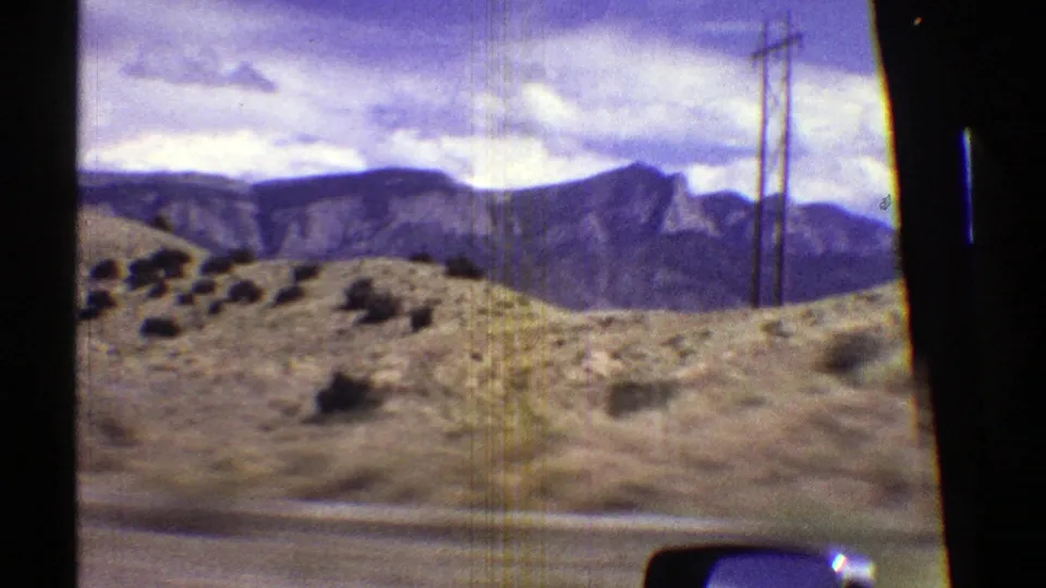 1969 Arizona road-trip frame with scrubby desert ground, roadside utility poles, and mountain ridges beyond the car window.