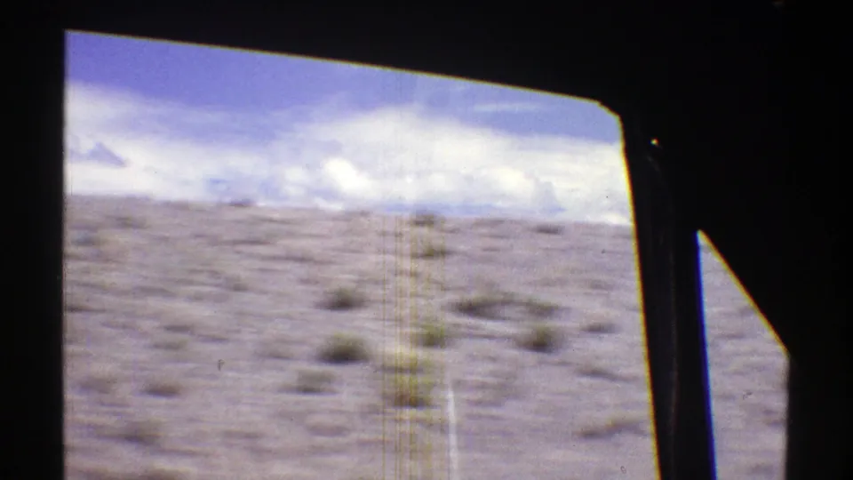 Open Arizona desert scrubland passes outside a dark vehicle window frame, with pale ground and clouded sky in soft 8mm color.