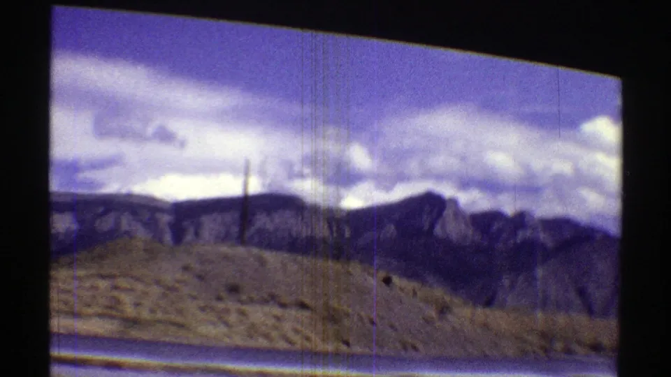 A 1969 Arizona road-trip frame shows scrubby desert ground, distant mountains, and a cloudy sky through a vehicle window.