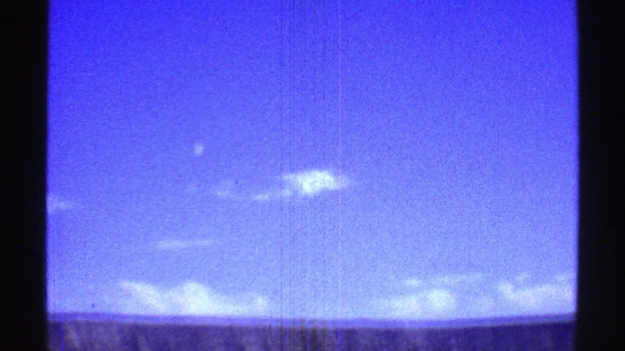 Blue sky with white clouds above a distant ridge, framed by dark 8mm film borders.