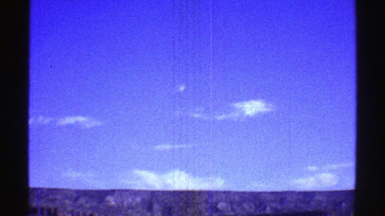 1969 Arizona landscape frame with blue sky, scattered white clouds, and a distant ridge seen through aged 8mm film texture.