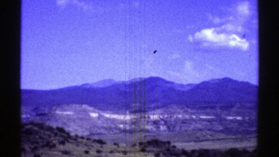 1969 Arizona home-movie still with a scrub-covered desert foreground, pale hills, and a dark mountain range under a clear blue sky with a small cloud.