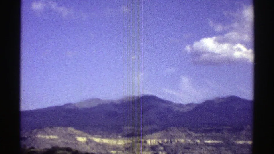 1969 Arizona 8mm frame showing a distant mountain ridge, pale desert foreground, blue sky, small clouds, and visible film scratches.