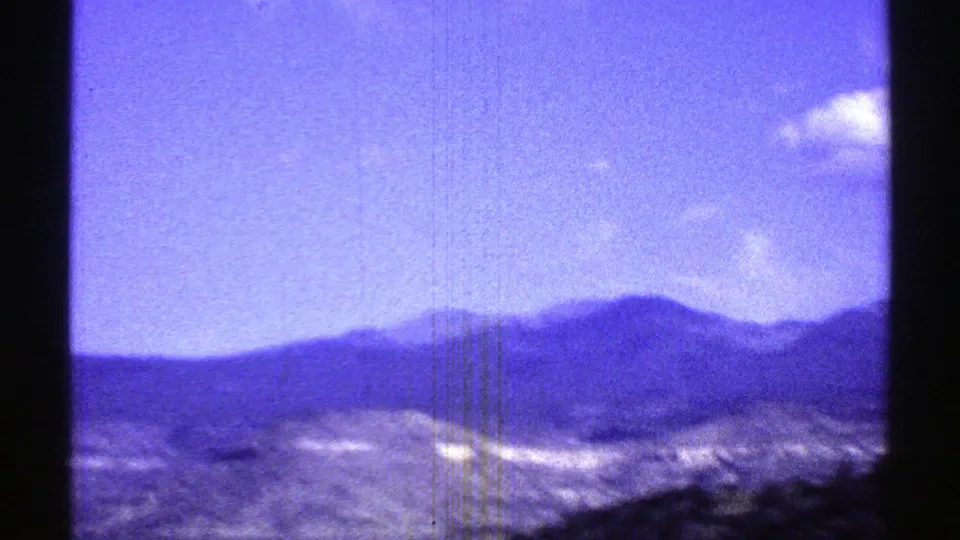 Soft 8mm Arizona frame showing a dark mountain ridge, rocky desert slope, open sky, and a small white cloud near the horizon.