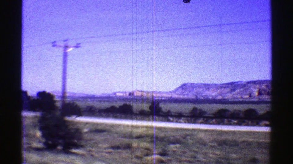 1969 Arizona 8mm roadside view with a utility pole, pale road edge, open desert fields, distant hills, and visible film wear.
