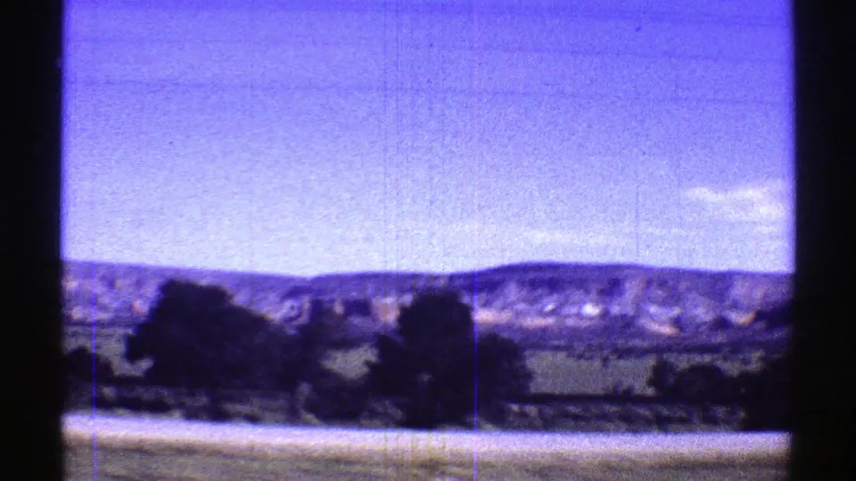1969 road-trip frame with dark roadside trees, green fields, distant hills, and motion blur from a moving vehicle.