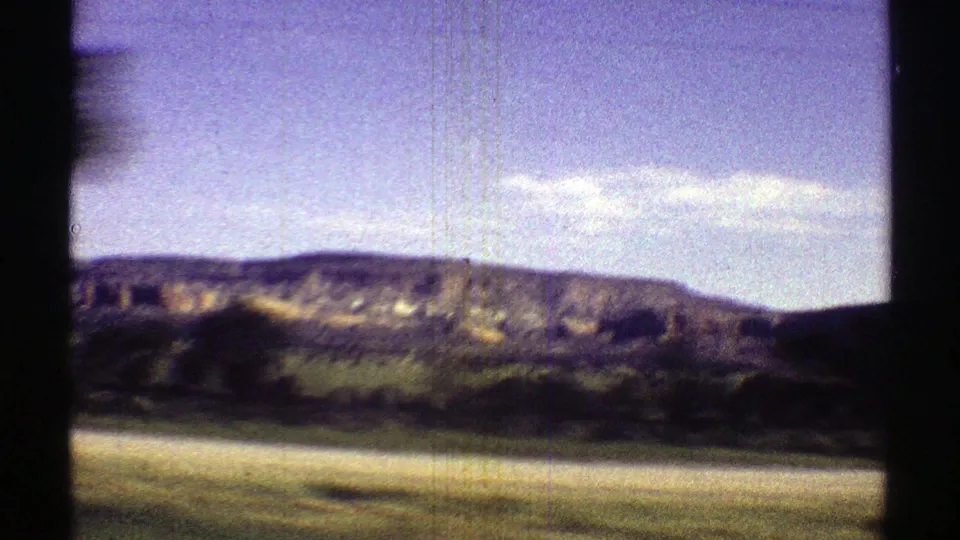 Arizona 8mm frame showing a low mesa beyond roadside fields, with strong daylight, grain, and slight color fading.
