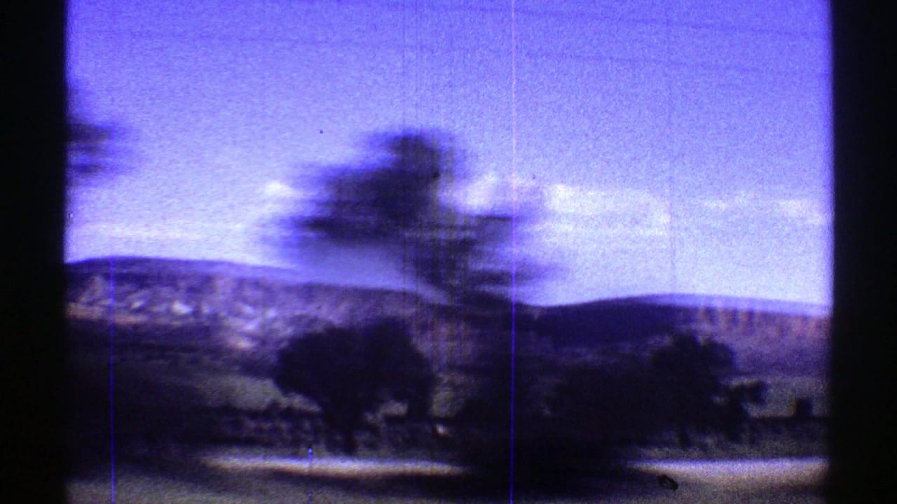 1969 Arizona home-movie frame showing blurred trees, low desert hills, and dark car-window edges during a moving roadside view.