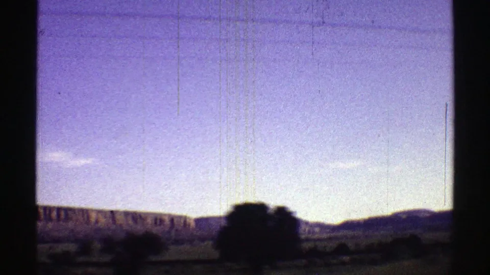 1969 Arizona road-trip frame with low desert ridges, dark foreground trees or brush, and a pale sky softened by 8mm exposure.