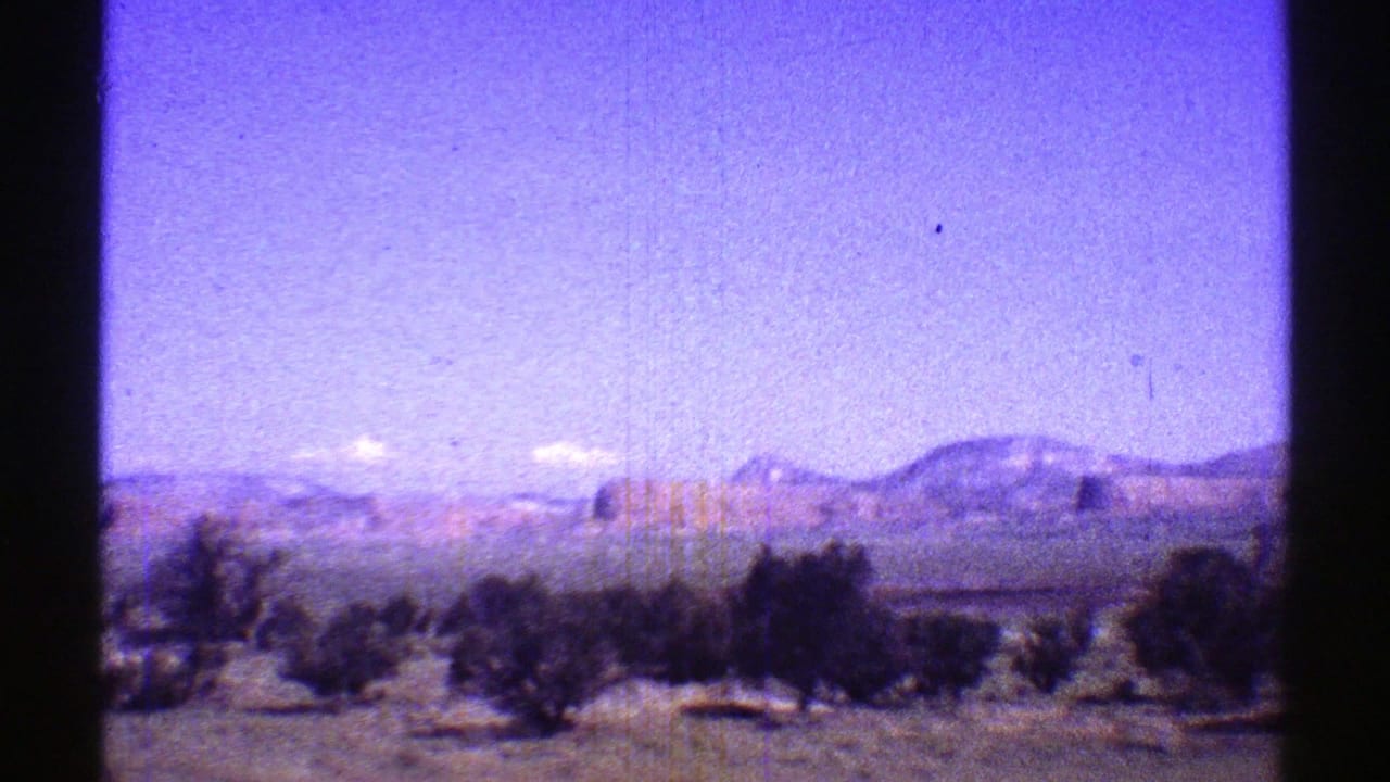 Faded 8mm frame of Arizona desert terrain with dark shrubs, distant mesas, and a color-shifted open sky.