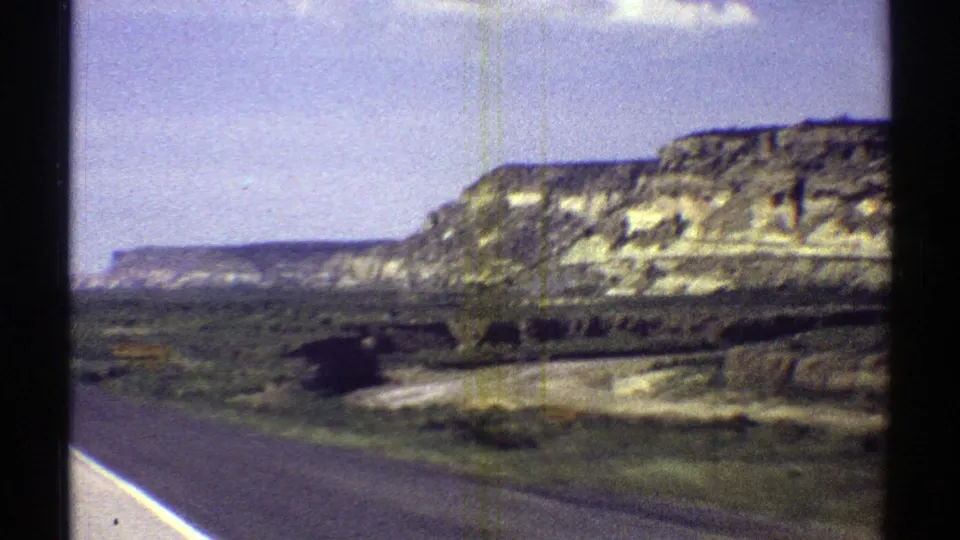 1969 Arizona roadside view with a paved road edge, green scrub, and layered mesa hills under a pale blue sky.