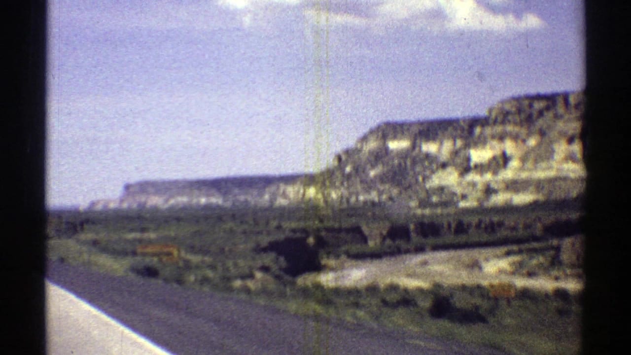1969 8mm Arizona road view with brown mesas, open scrub terrain, and a bright blue sky with small clouds.
