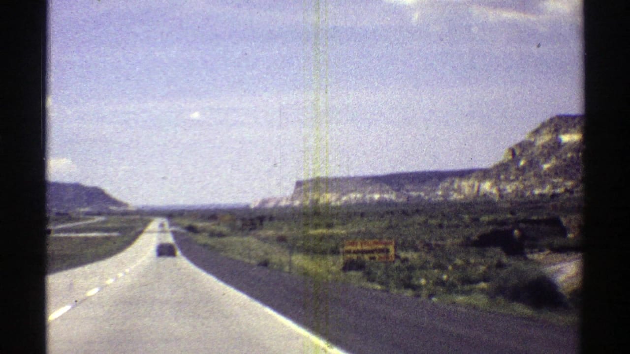 Arizona desert road stretches ahead in 1969 8mm footage, with a distant vehicle and mesas on both sides.