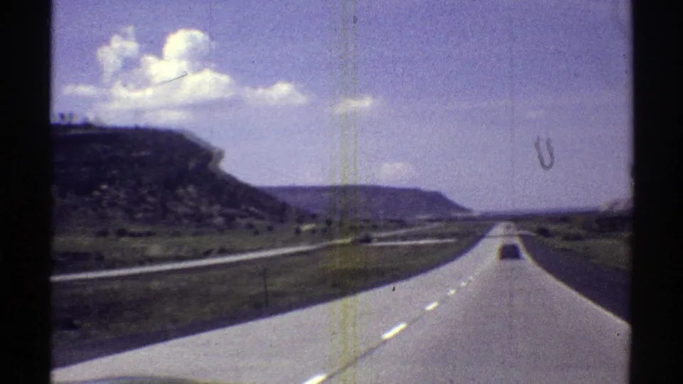 Forward-facing 8mm view of an Arizona highway, distant vehicle, mesa cliffs, and white clouds in a blue sky.